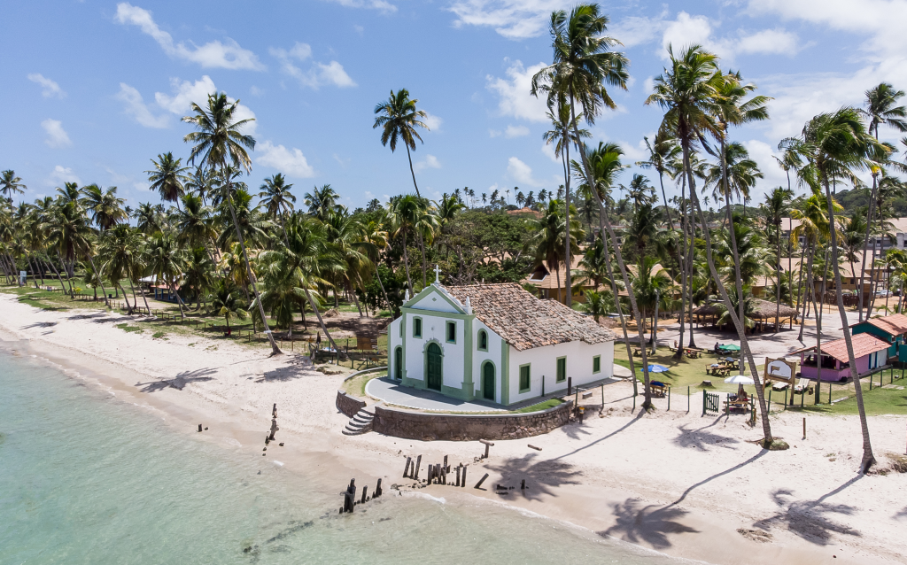 Vista aérea de uma praia tropical com palmeiras e uma charmosa construção branca com detalhes verdes, rodeada por areia e natureza.