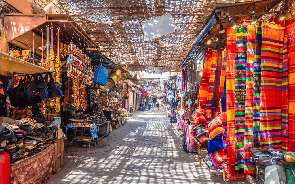 Rua estreita no souk de Marrakech, Marrocos, com barracas vendendo sapatos, tapetes e outros produtos tradicionais.