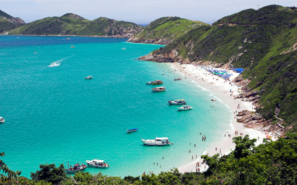 Praia paradisíaca com águas cristalinas e areia branca, rodeada por montanhas verdes e barcos ancorados, ideal para relaxamento e lazer.