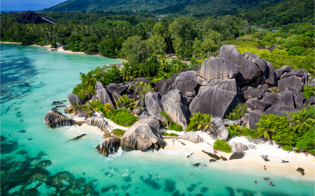 Vista aérea de uma praia nas Seychelles, com grandes formações rochosas de granito, águas turquesas e vegetação tropical ao fundo.