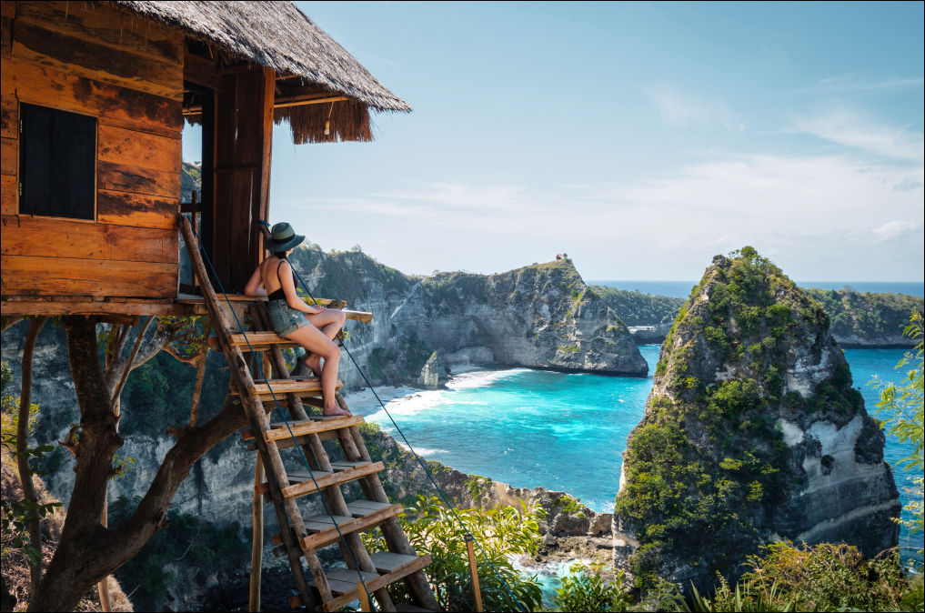 Uma mulher admira a vista do oceano a partir de uma casa na árvore. A paisagem é de tirar o fôlego, com águas cristalinas e formações rochosas impressionantes.