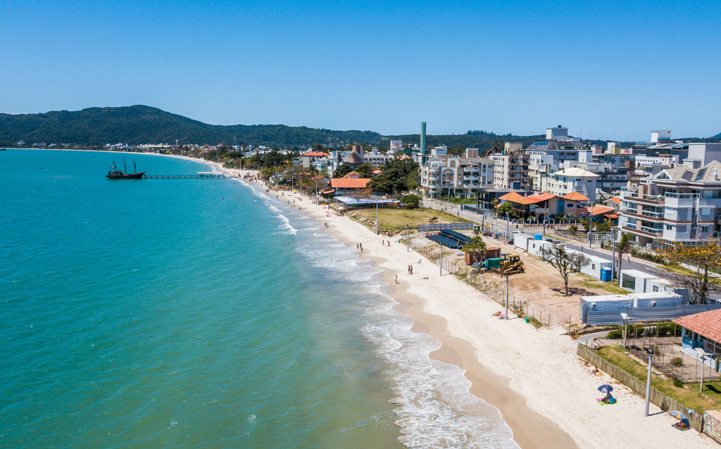 Praia com águas claras e areias brancas, cercada por construções modernas e natureza preservada, ideal para lazer e turismo.