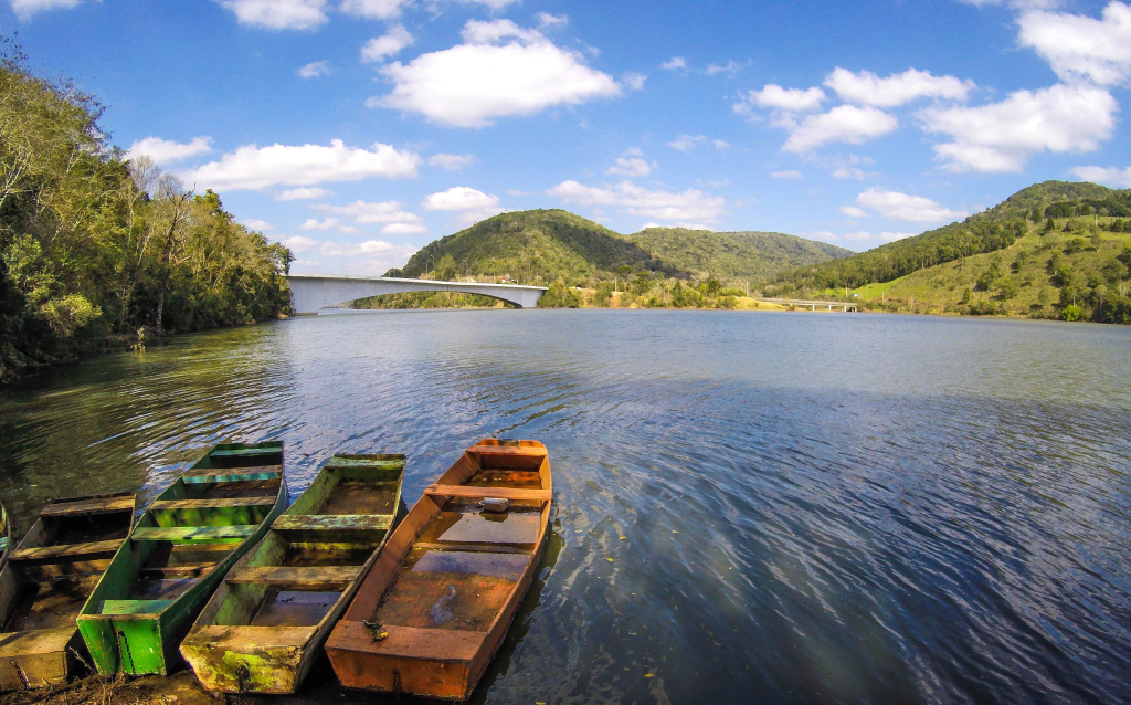 Imagem de um lago sereno com barcos de madeira coloridos na margem, cercado por colinas verdes e céu azul com nuvens brancas. Um ótimo lugar para relaxar e apreciar a natureza.