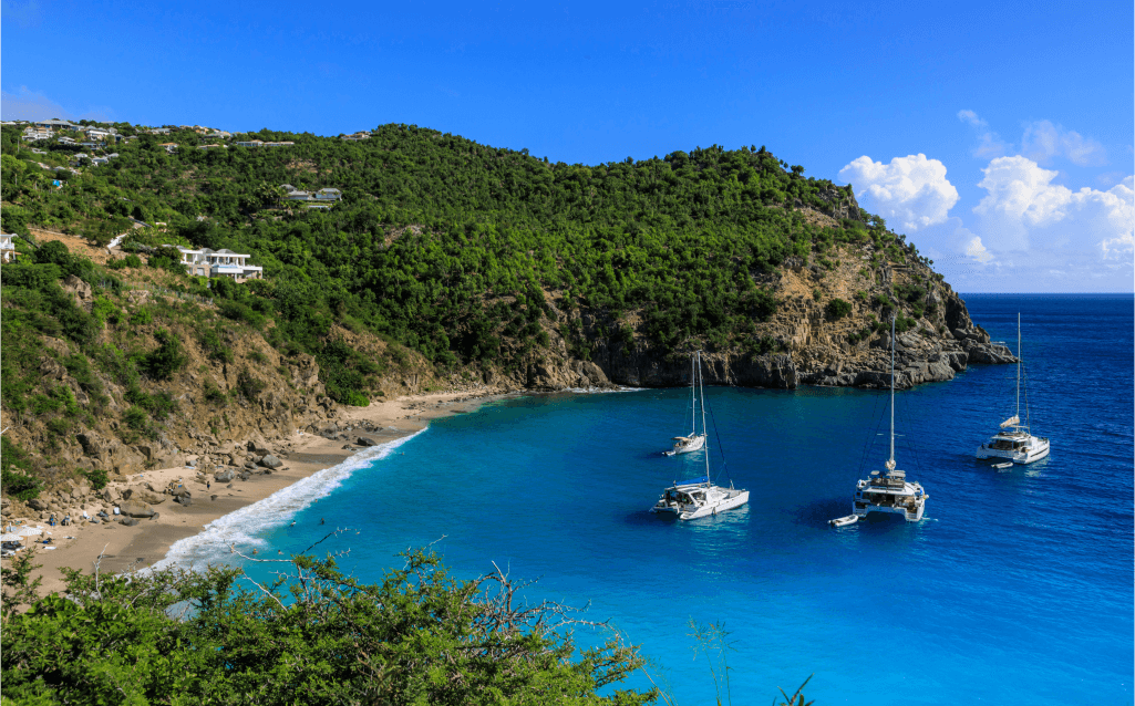 Enseada tranquila em Saint Barthélemy, com águas azuis e transparentes. Barcos ancorados flutuam próximo à praia isolada, cercada por colinas verdes sob um céu claro.