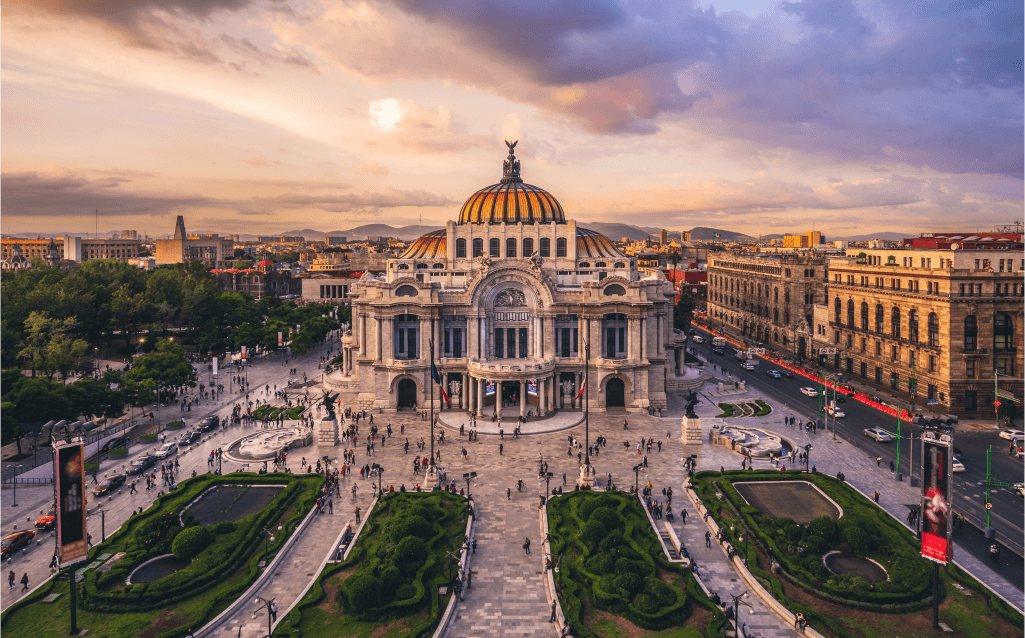 Vista aérea do Palácio de Bellas Artes na Cidade do México, com jardins ao redor e montanhas ao fundo.