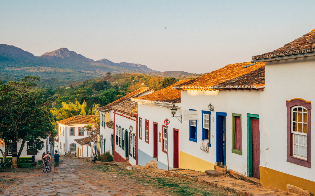 Vista de uma rua histórica com casas coloridas em uma colina, cercada pela natureza e montanhas ao fundo, simbolizando a cultura e turismo.