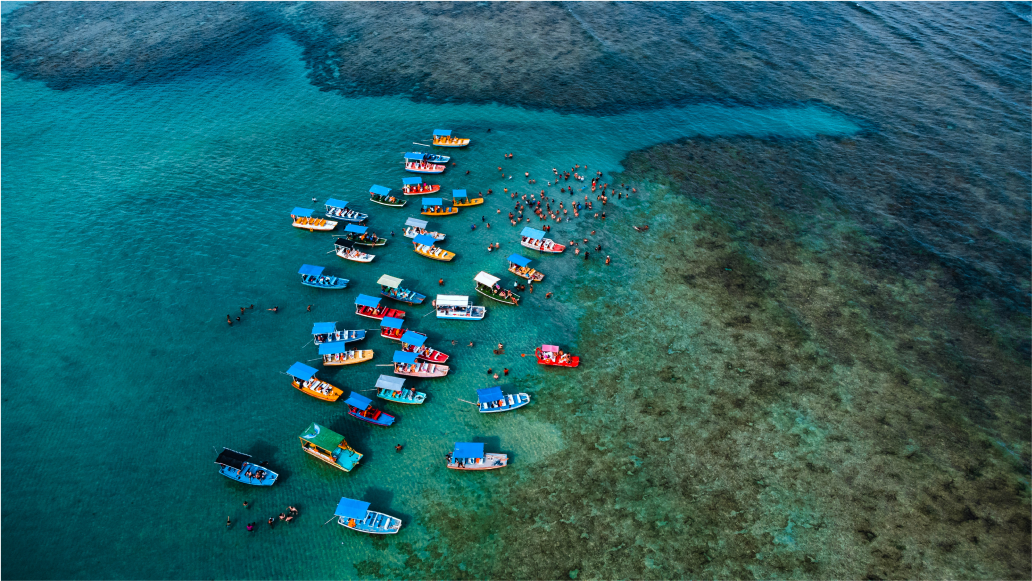 Vista aérea de uma praia com várias embarcações, na região tropical, destacando a beleza natural da costa e a urbanização ao fundo.