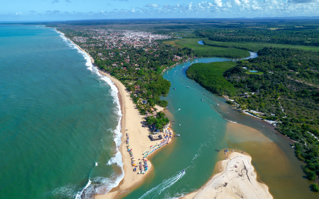 Vista aérea de uma bela praia cercada pela vegetação, com ondas do mar e um rio que desagua em sua extensão, ideal para turismo e lazer.