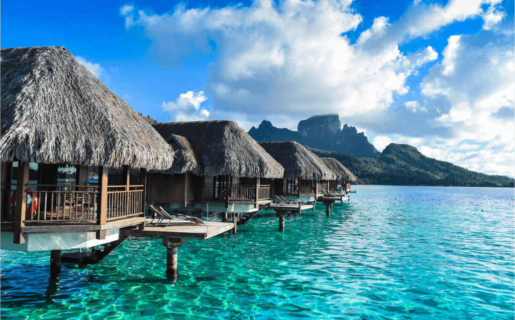 Bangalôs sobre águas azul-turquesa em Bora Bora, Polinésia Francesa, com montanhas ao fundo e céu parcialmente nublado, em um cenário de luxo e tranquilidade.