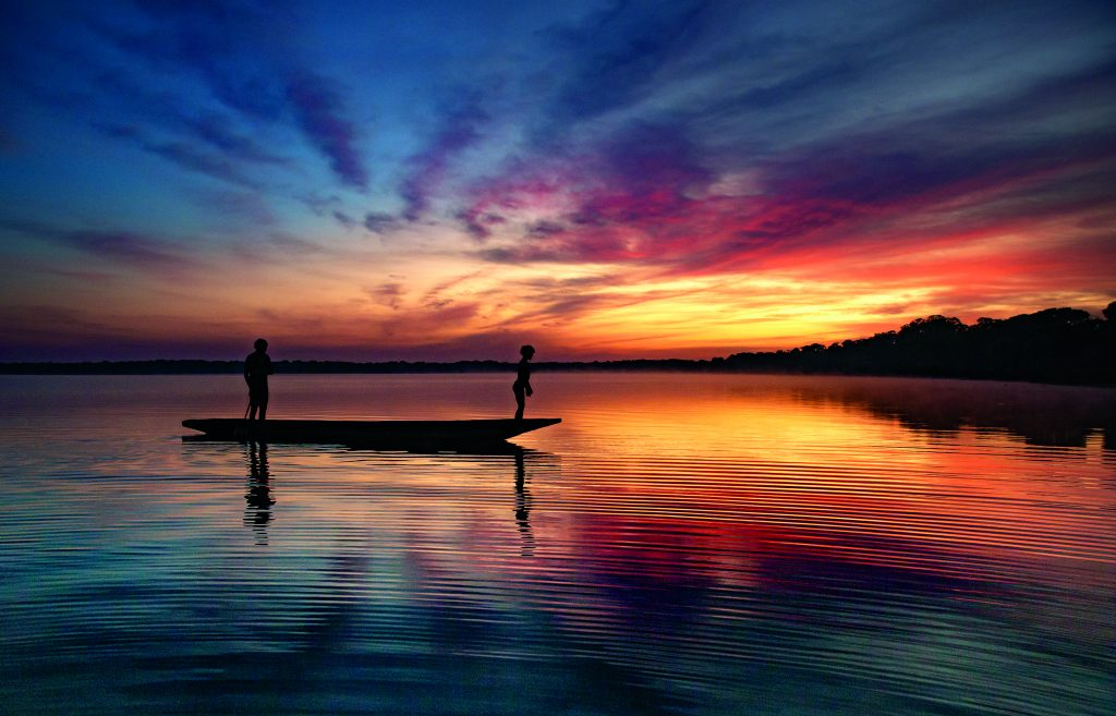 Foto de uma pequena canoa onde se pode ver a silhueta de duas pessoas em pé sobre ela, sendo pelo menos uma delas uma criança. No fundo, o pôr do sol e o reflexo no rio nas cores amarelo, laranja, rosa, roxo, azul e vermelho.