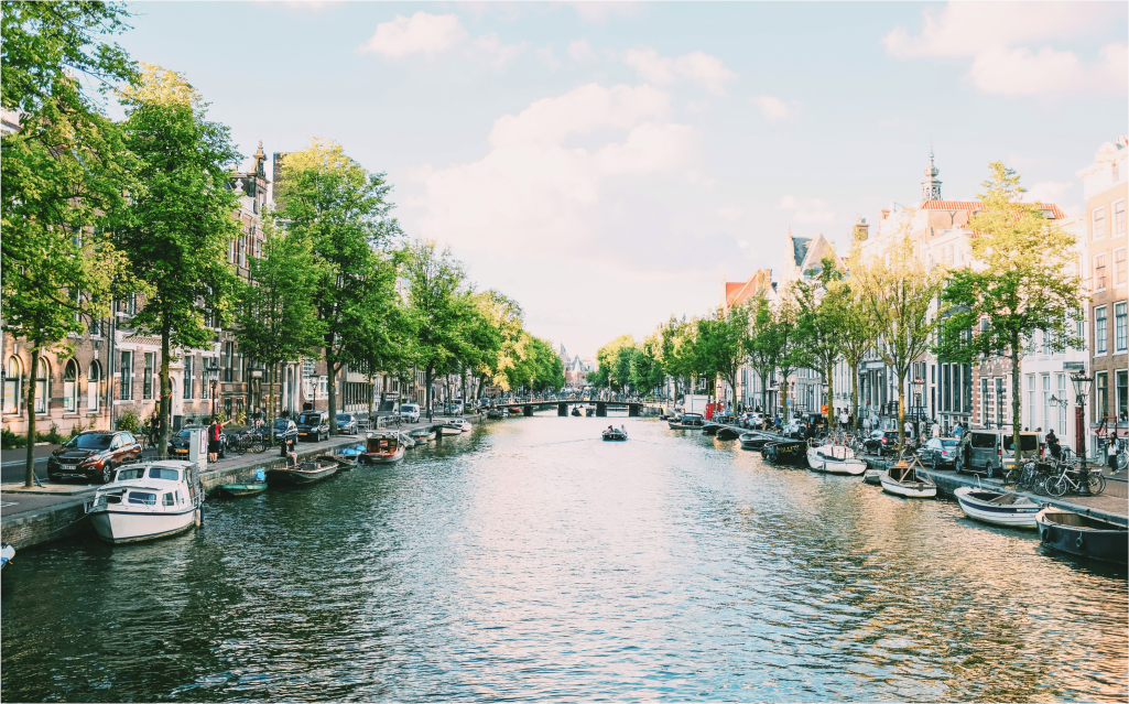 Vista encantadora de um canal em Amsterdam, com árvores verdes ao longo das margens, barcos à deriva e edifícios típicos da arquitetura holandesa ao fundo. Perfeito para quem procura beleza e tranquilidade.