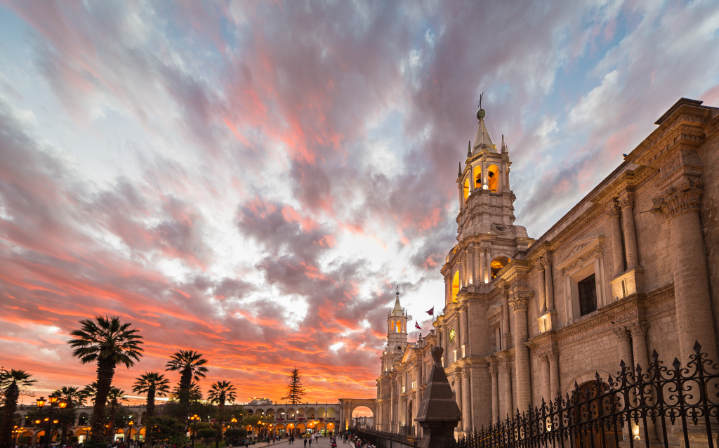   A bela arquitetura da cidade de Arequipa, Peru, ao entardecer, destacando as nuvens coloridas no céu e palmeiras ao longo da calçada.