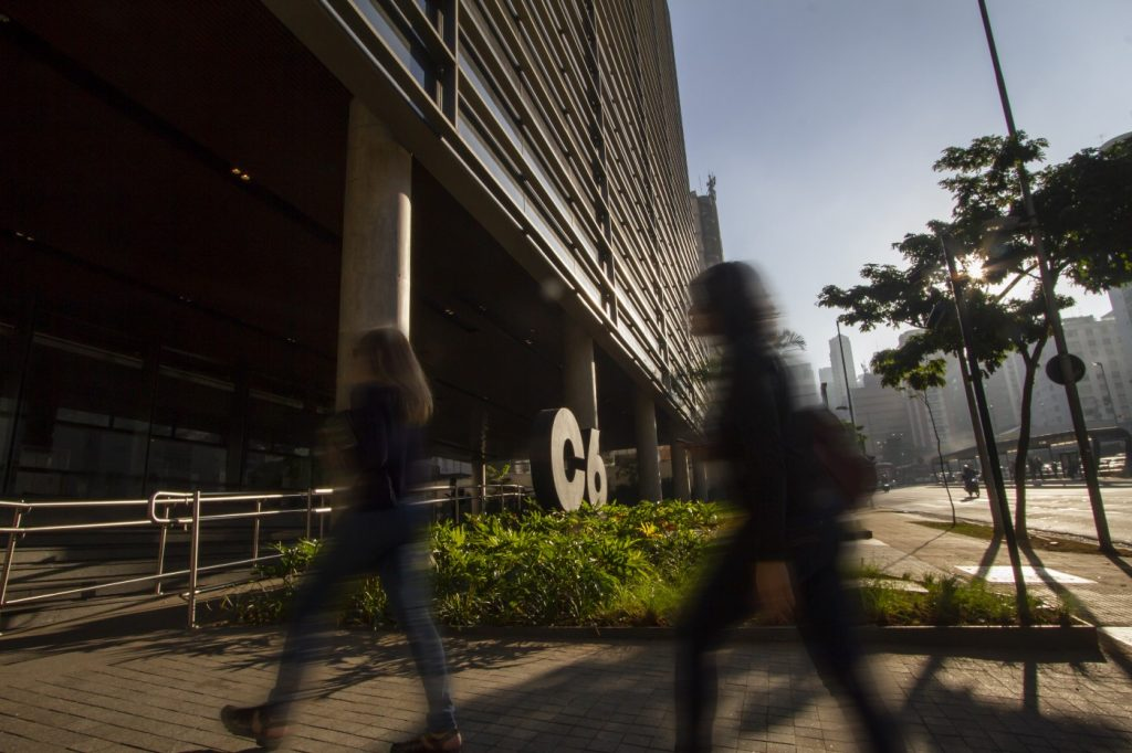 Foto da entrada do C6 Bank, com duas pessoas passando em primeiro plano no sentido da entrada do banco. No meio dela, vemos o logo do C6 Bank colocado no jardim, refletindo a luz do sol.