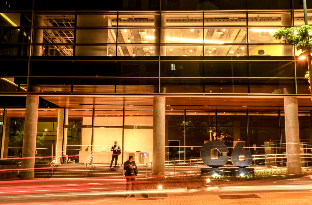 Foto da fachada do prédio do C6 Bank, onde se pode ver movimento do carro em time lapse e duas pessoas mexendo no celular