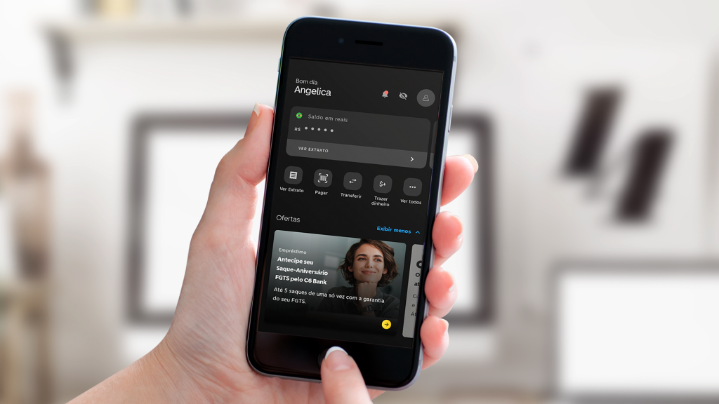 Foto da mão esquerda de uma mulher branca segurando um celular com a tela inicial do app do C6 Bank aberta