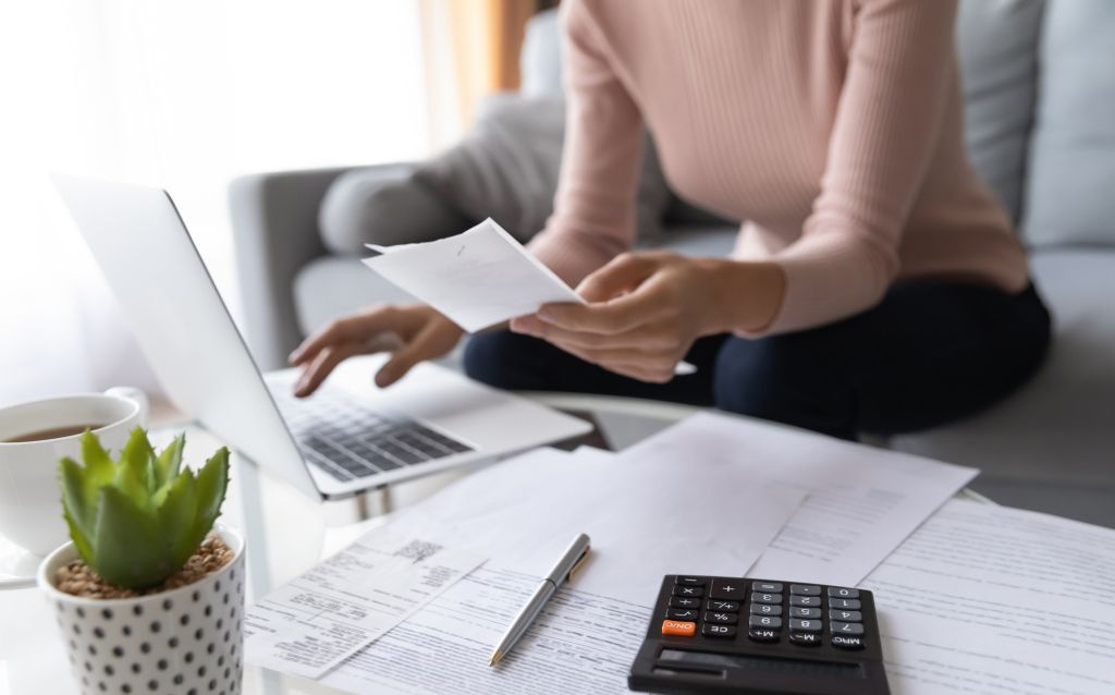 como a população sente a variação do poder de compra  | mulher sentada na sala mexendo em um notebook e segurando um papel, na mesa em sua frente tem um cacto, papéis e calculadora