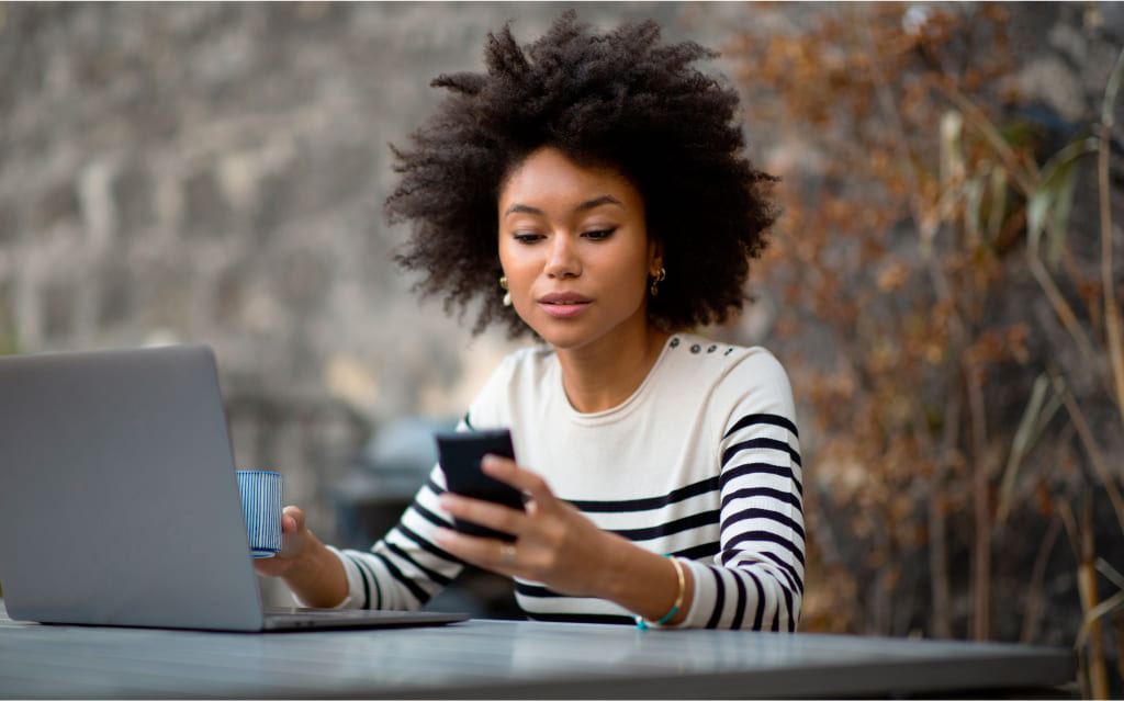 mulher negra sentada diante de computador segurando celular na mão esquerda e pesquisando como calcular cdb