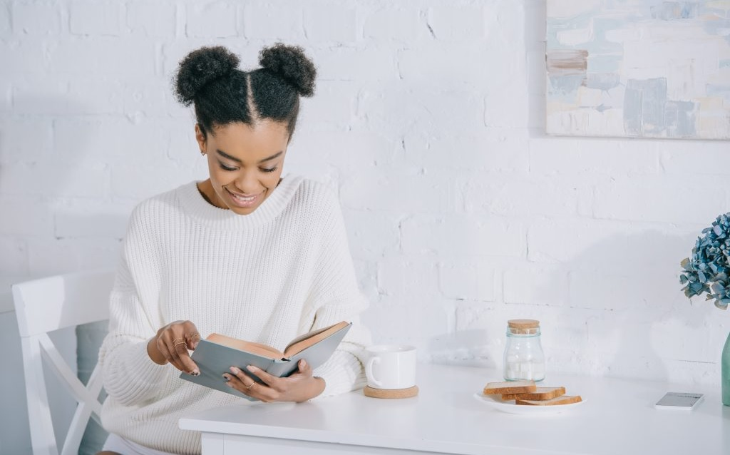 mulher negra usando roupas brancas sentada sorri enquanto lê livro sobre dívida boa