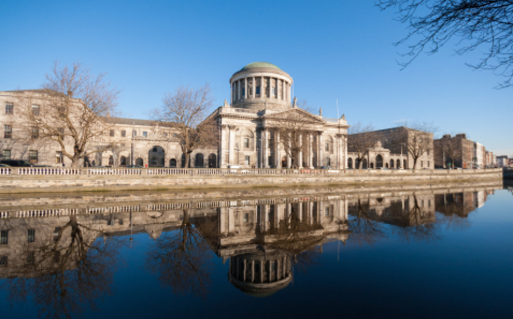 A imagem mostra o elegante edifício do Antigo Tribunal da Cidade de Dublin, refletido nas águas calmas do rio. Em um dia ensolarado, a arquitetura neoclássica destaca-se sob o céu azul, iluminando a história da cidade.