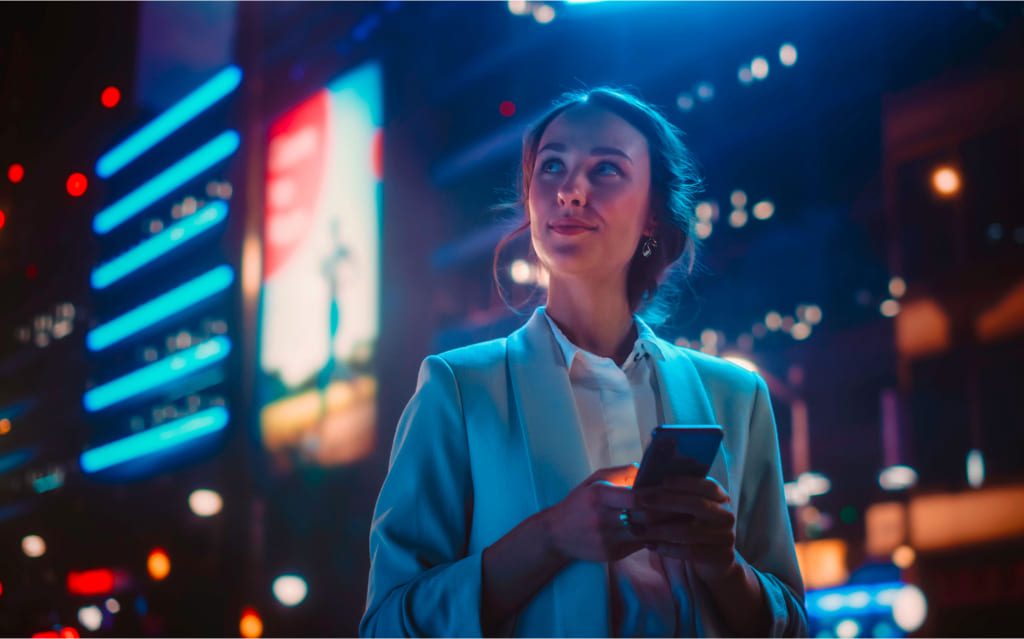 mulher à frente de prédios luminosos olhando para o alto e segurando celular nas mãos após pesquisar fundos multimercado como funciona