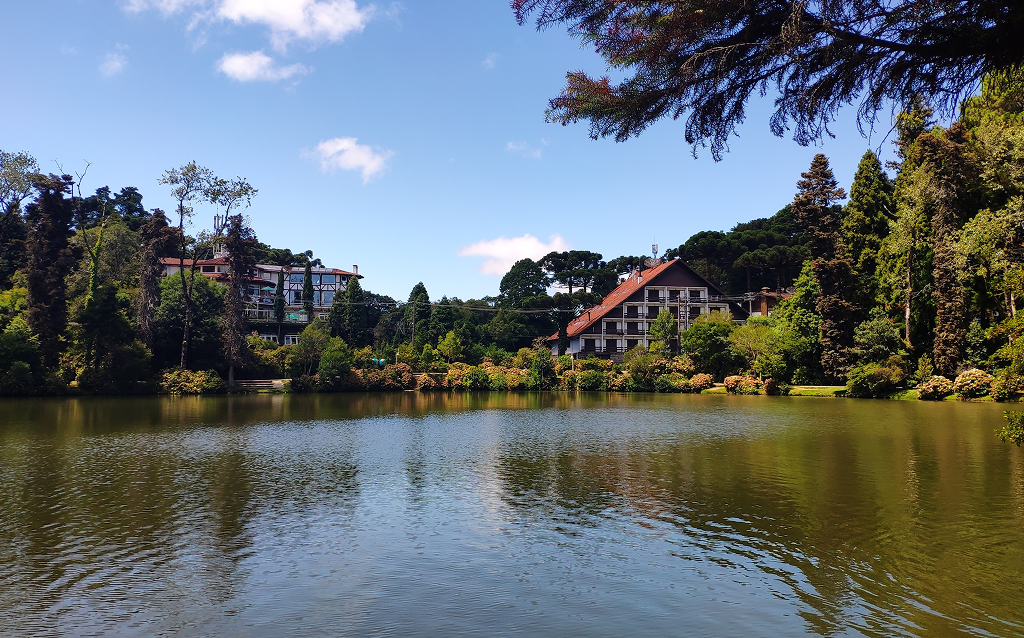 Vista de um lago rodeado por natureza exuberante e edificações charmosas em um dia ensolarado. A tranquilidade do local convida ao descanso e ao lazer.