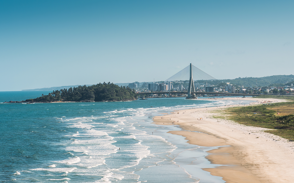 Vista panorâmica de uma linda praia com ondas quebrando, cercada por uma cidade e uma ponte moderna ao fundo, ideal para turismo e relaxamento.