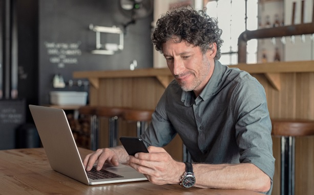 homem grisalho usando camisa social sentado de frente para computador com celular nas mãos pesquisando o que significa risco arrojado