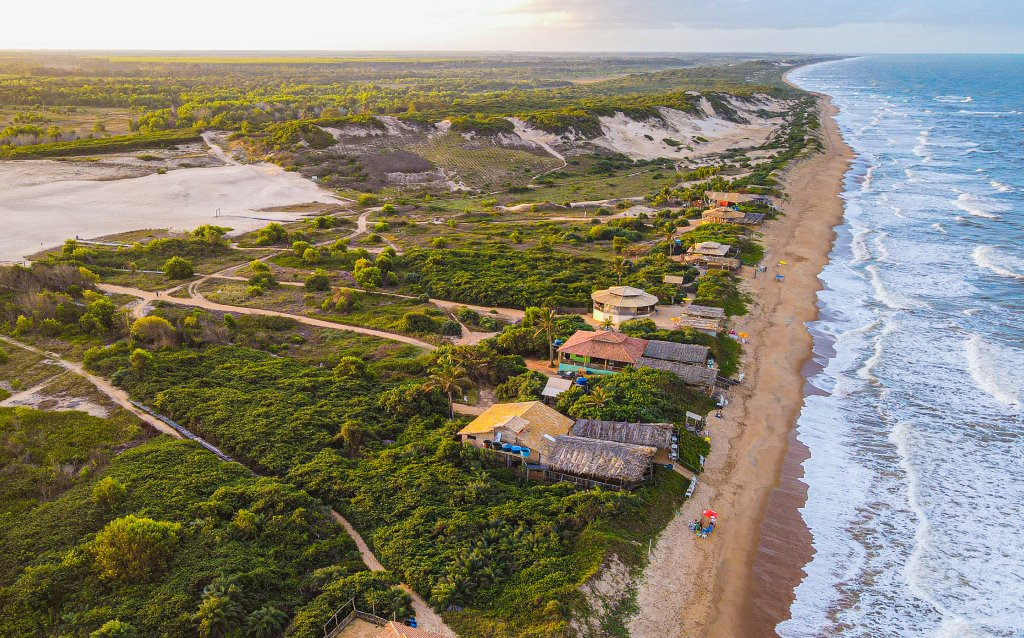 Vista de cima da cidade de Itaúnas. Do lado direito está a areia e o mar, e do esquerdo, as casas de praia, dunas e diversas árvores que completam as paisagens.