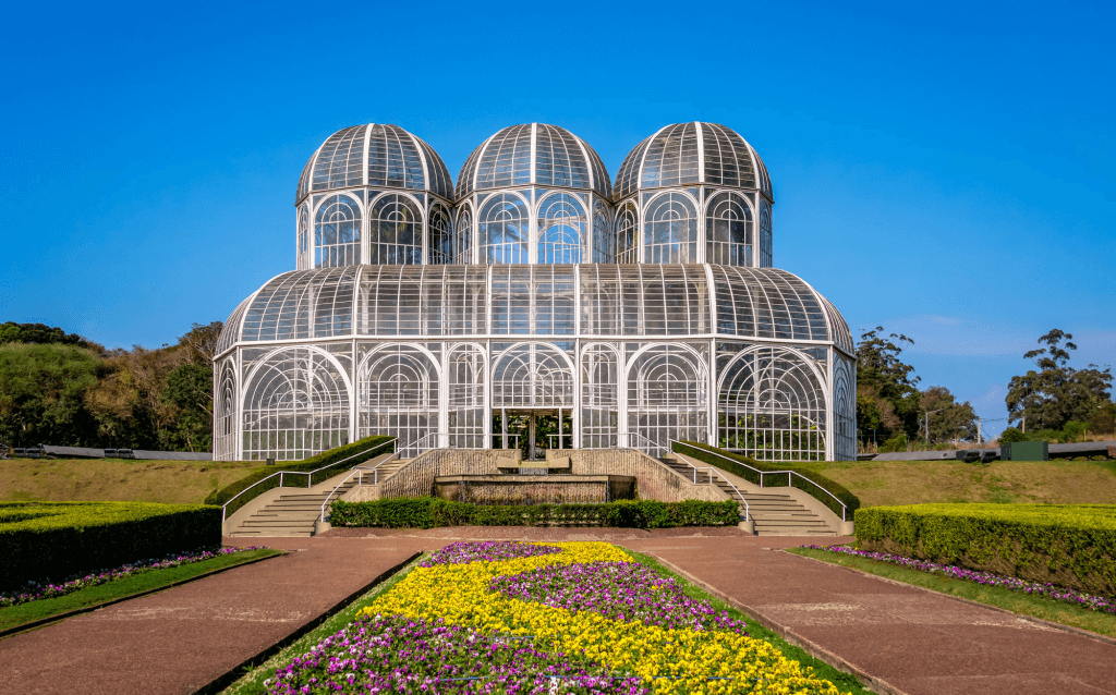 Jardim Botânico de Curitiba: Paisagem serena destacando os canteiros floridos e a arquitetura singular da estufa. Uma variedade de flores coloridas emoldura o cenário, criando uma atmosfera tranquila e natural.