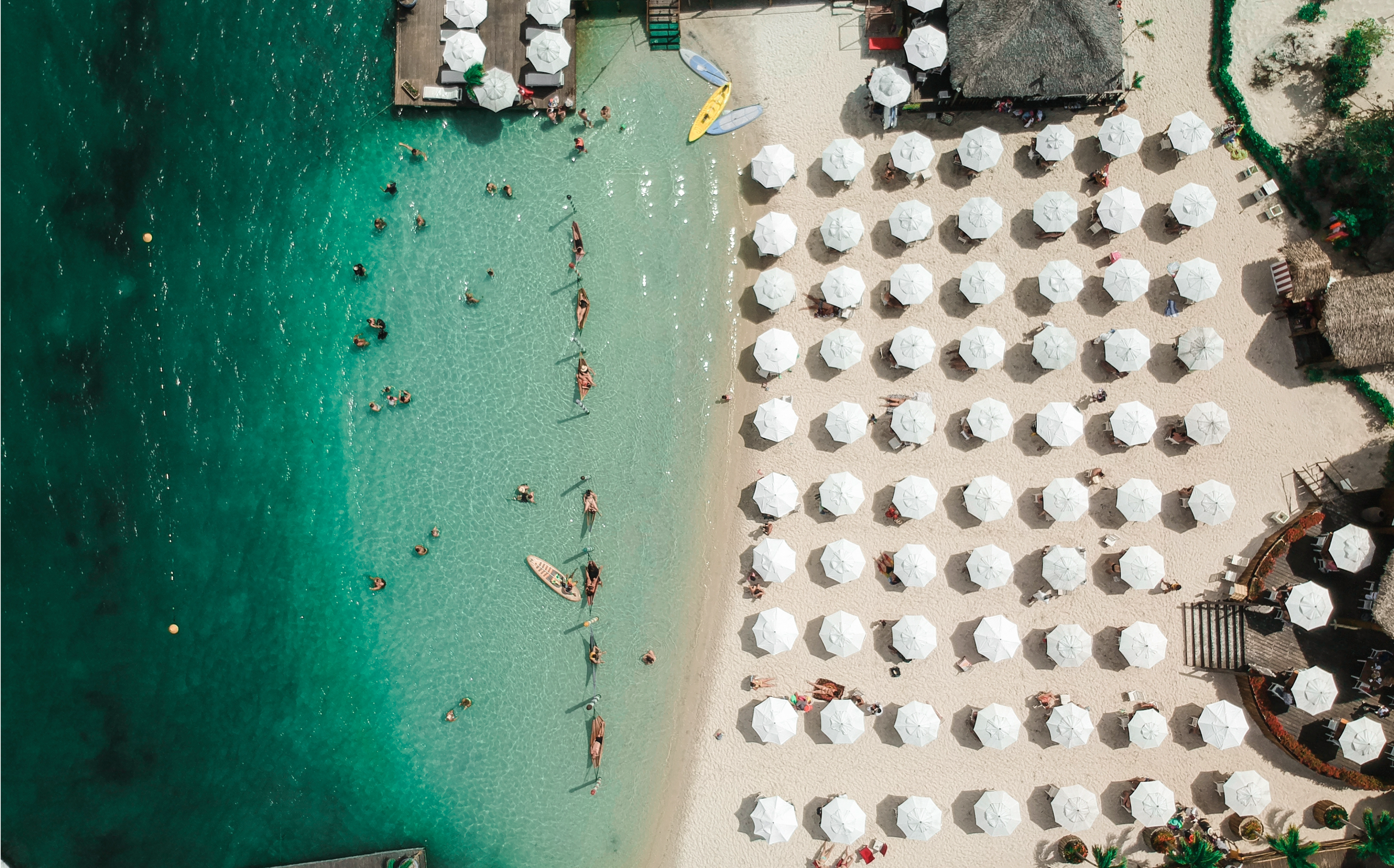 Imagem aérea de uma praia paradisíaca com águas cristalinas, várias pessoas se divertindo na água e muitas cadeiras com guarda-sóis brancos na areia.