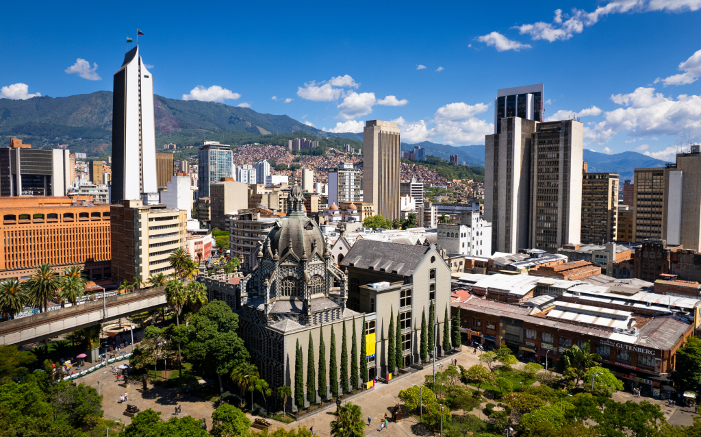 Vista panorâmica da cidade de Medellín, com arranha-céus, montanhas ao fundo e a arquitetura da Igreja de San Pedro. Um contraste entre o moderno e o histórico.