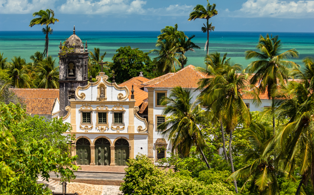 Vista panorâmica de uma construção histórica com arquitetura colonial, rodeada por palmeiras e com o mar azul ao fundo, típica do Brasil.