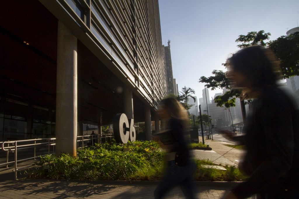 Foto da fachada do prédio do C6 Bank durante um dia de sol, onde se pode ver o vulto de duas pessoas em movimento entrando no banco e o logo do C6 Bank no jardim ao fundo.