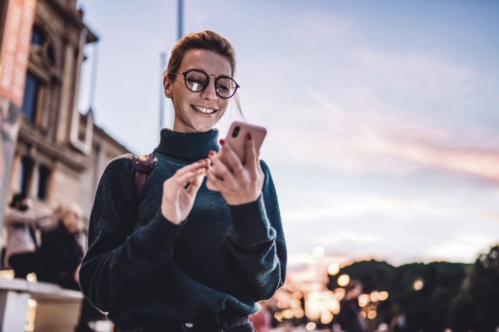 Foto de uma mulher loira com uma casaco verde militar e óculos grandes lilás mexendo em um smartphone com capinha rosa e sorrindo. No fundo é possível ver o por do sol e o céu e tons de lilás