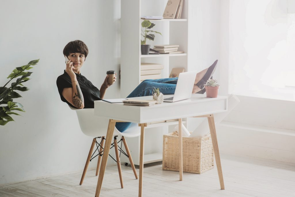 Foto de uma jovem branca, morena, com o cabelo curto no estilo channel. Ela está sentada numa cadeira com os dois pés apoiados em cima de uma mesa a sua frente, segurando um celular na orelha com a mão direita e um café com a mão esquerda. Na cena ainda vemos um notebook e alguns papéis em cima da mesa e uma estante ao fundo. Ela está entrando em contato com o chat da C6 Store