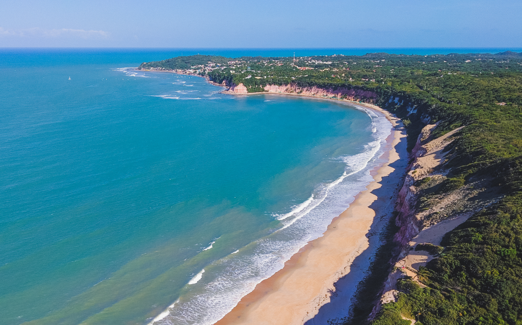 Vista aérea de uma paisagem costeira com penhascos, uma praia de areia e ondas azul-turquesa sob um céu azul claro.