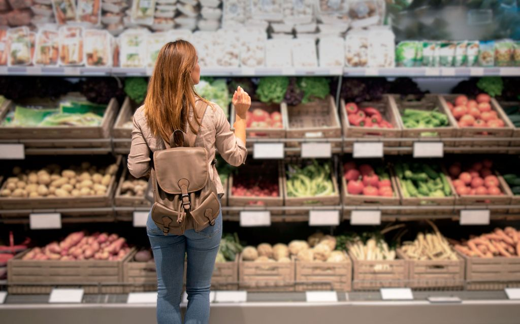 Mulher no mercado olhando para diferentes tipos de frutas e vegetais, analisando o preço abusivo dos produtos