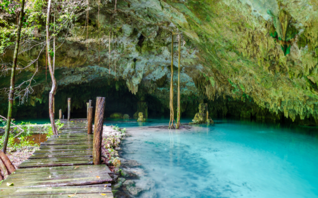 Cenário natural com uma caverna repleta de estalactites, um riacho de água azul clara e uma passarela de madeira no meio da vegetação tropical.