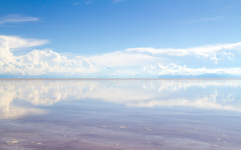 Imagem de um vasto horizonte refletindo águas calmas sob um céu azul claro e nuvens brancas, representando um cenário tranquilo e sereno na natureza.