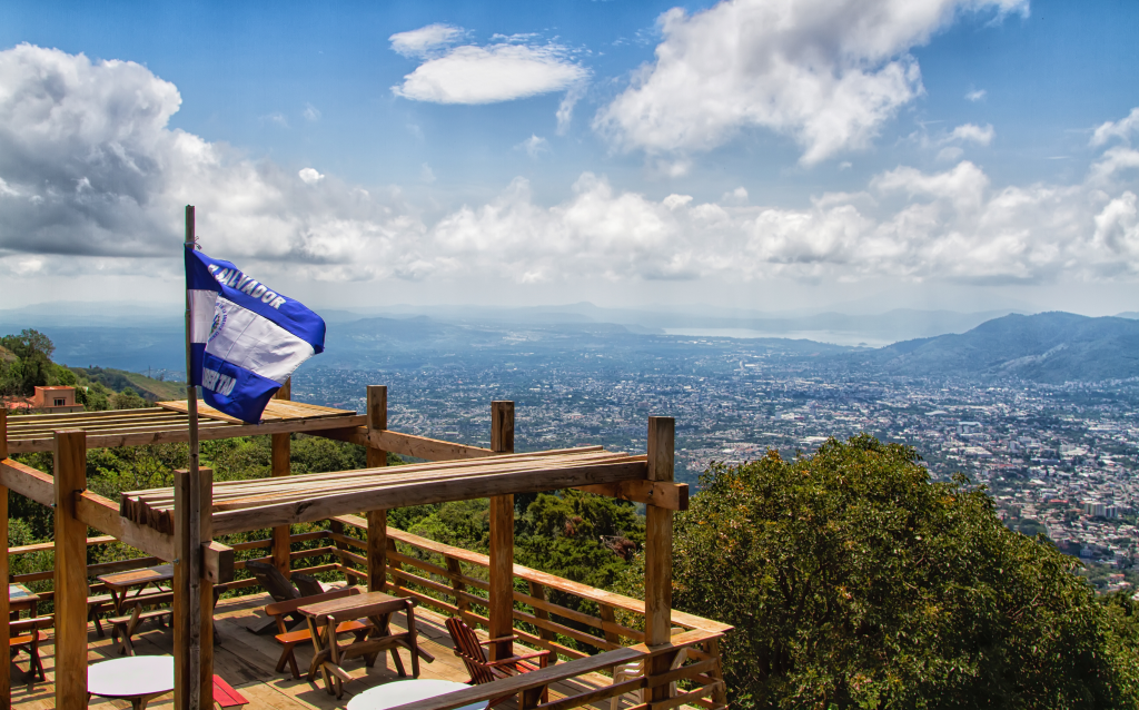 Vista panorâmica de uma cidade dominada por montanhas, com uma bandeira ao vento e um deck de madeira. O céu está parcialmente nublado, criando um contraste bonito.