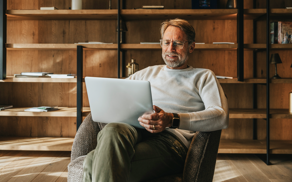   Homem sorridente usando laptop em ambiente aconchegante, com estante ao fundo, perfeito para trabalhar ou relaxar em casa.