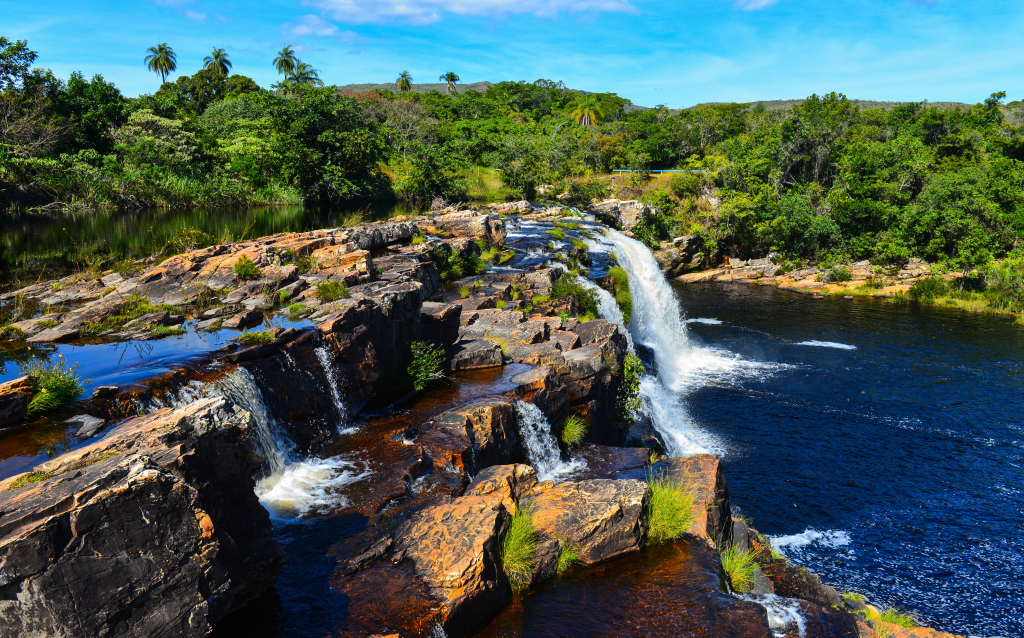 Belíssima cachoeira em meio à natureza exuberante, com rochas e vegetação ao redor, refletindo no rio tranquilo.