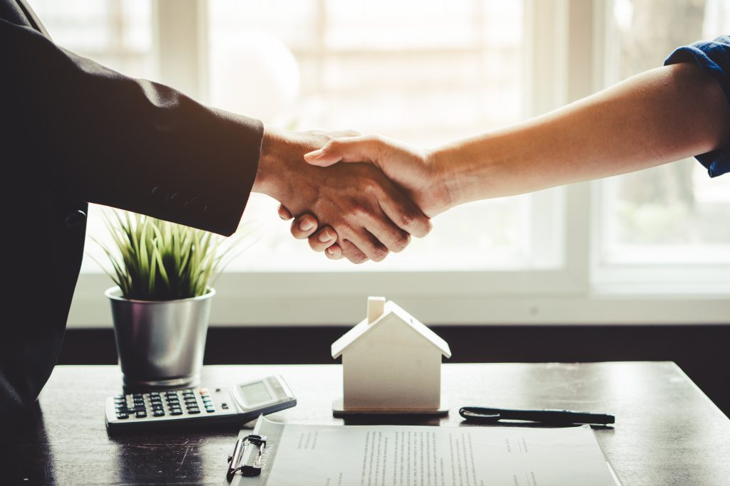 Duas pessoas dando as mãos, para fechar um negócio. Uma casinha, uma calculadora e papeis estão em cima de uma mesa