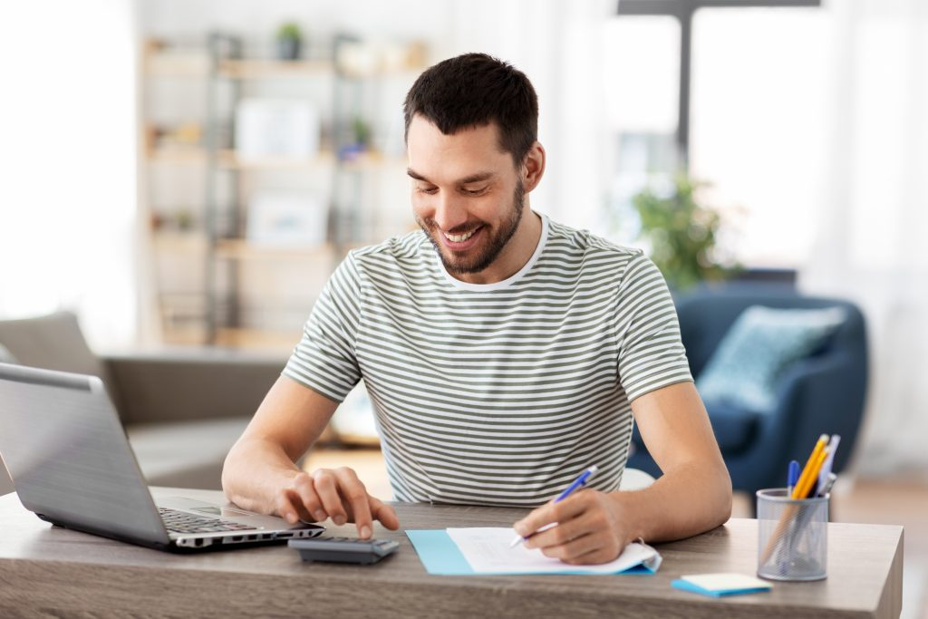 Homem branco de camiseta listrada de preto e branco, sentado em uma mesa, usando a calculadora e sorrindo