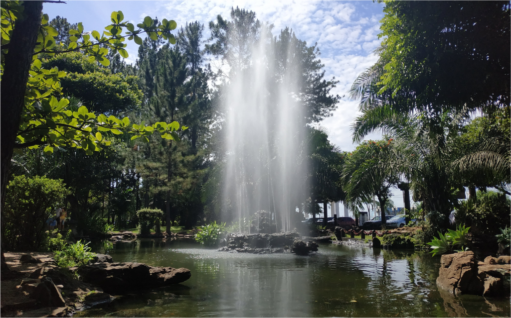 Uma bela fonte jorrando água em um lago rodeado por árvores e plantas exuberantes, sob um céu azul com nuvens