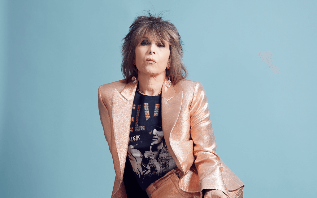 Chrissie Hynde, vocalista dos The Pretenders, veste blazer rosa metálico e camiseta preta, posando contra um fundo azul claro.