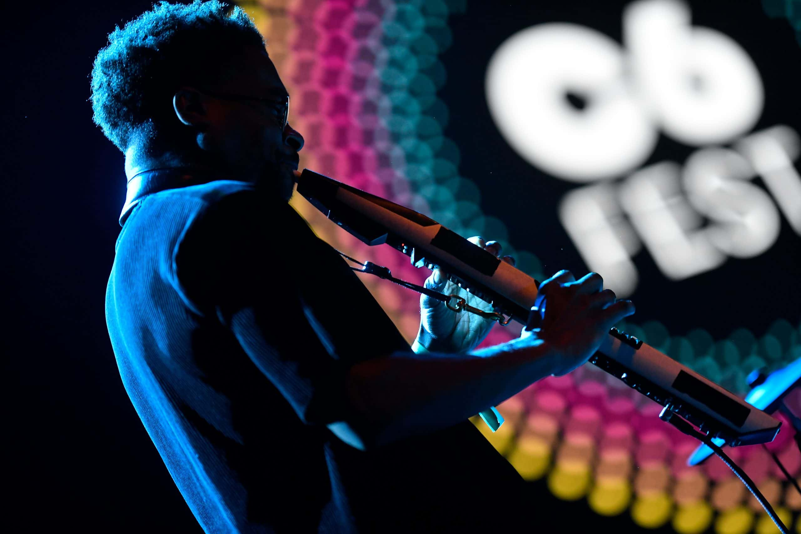 A imagem mostra um instrumentista no primeiro plano de imagem. Ele está de costas e sendo iluminado por uma luz azul. Ao fundo, está o logo do C6 Fest desfocado.