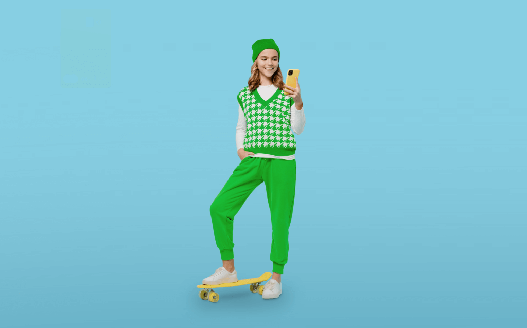 Menina de pele branca, usando uma touca, calça e colete verdes e um tênis branco. Segura um celular de capa amarela e seu pé esquerdo está em cima de um skate também amarelo. O fundo da imagem é azul.