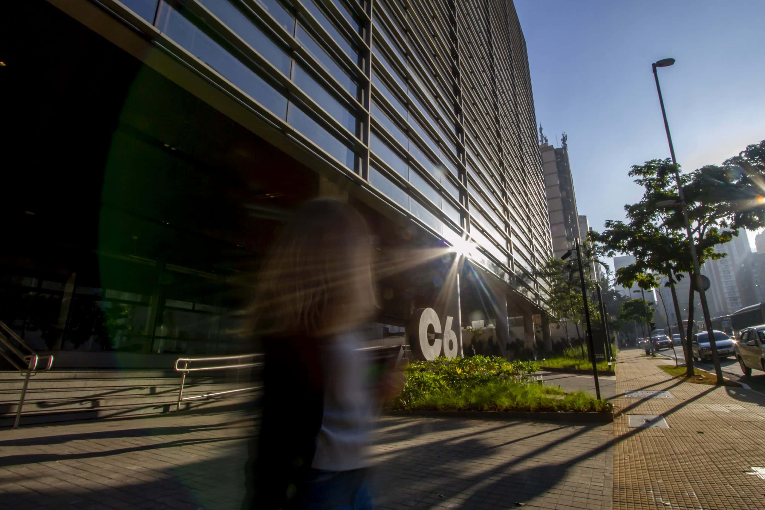 Foto do prédio do C6 Bank na Avenida Nove de Julho, com uma pessoa olhando na direção do logo do banco e da luz do sol refletindo no vidro da fachada do banco.