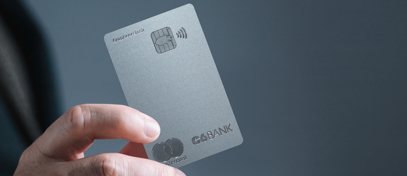 Foto da mão de uma pessoa branca segurando o cartão C6 Graphene, um cartão feito de axo inoxidável com a cor predominantemente prateada, com o logo do C6 Bank e da Mastercard, o símboldo da tecnologia NFC e a inscrição Graphene Black na parte frontal do cartão.